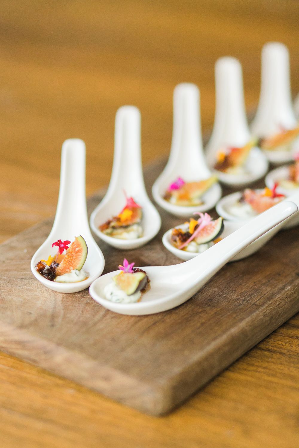 A wooden serving board with white spoons containing food garnished with flowers.