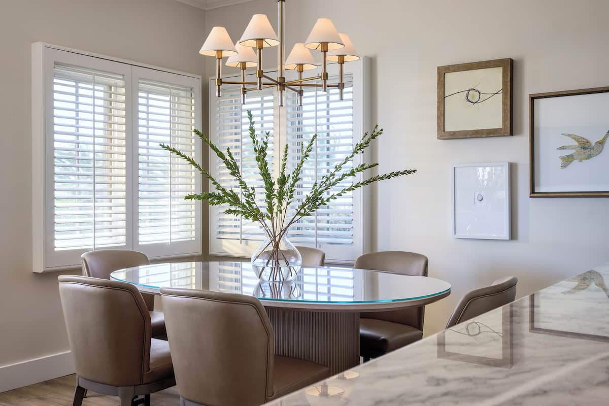A dining area with a round glass table, beige chairs, a vase with greenery, and a chandelier.