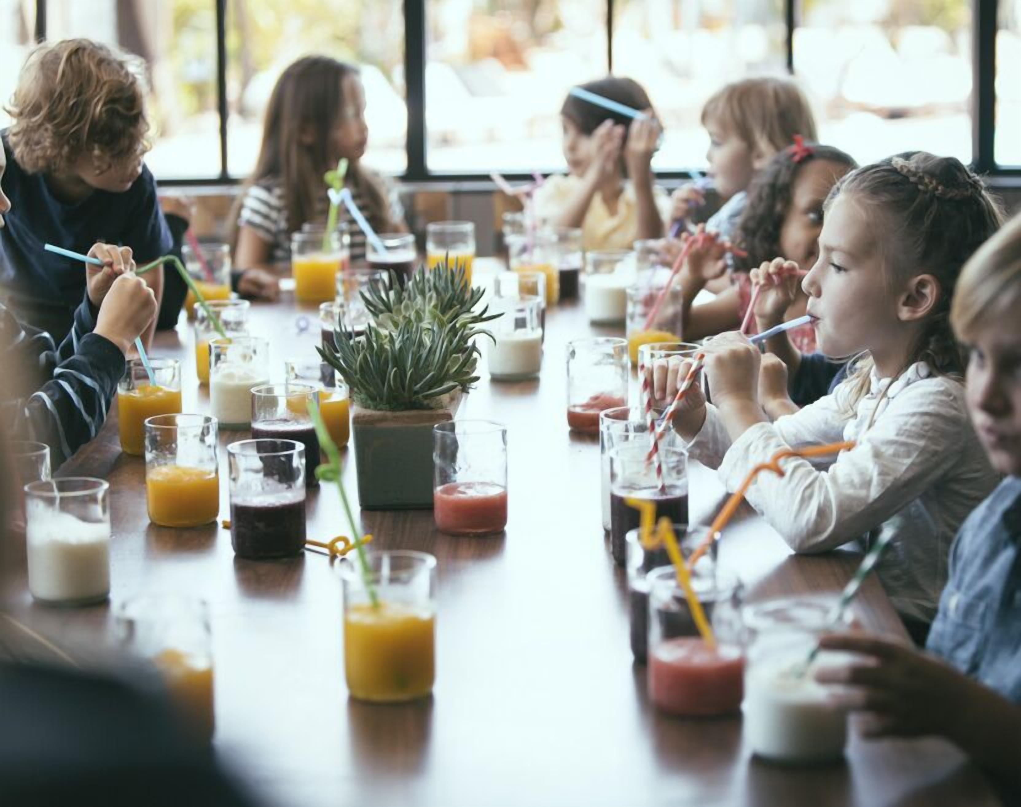 Un grupo de niños sentados a una mesa con diversas bebidas y una planta en maceta.