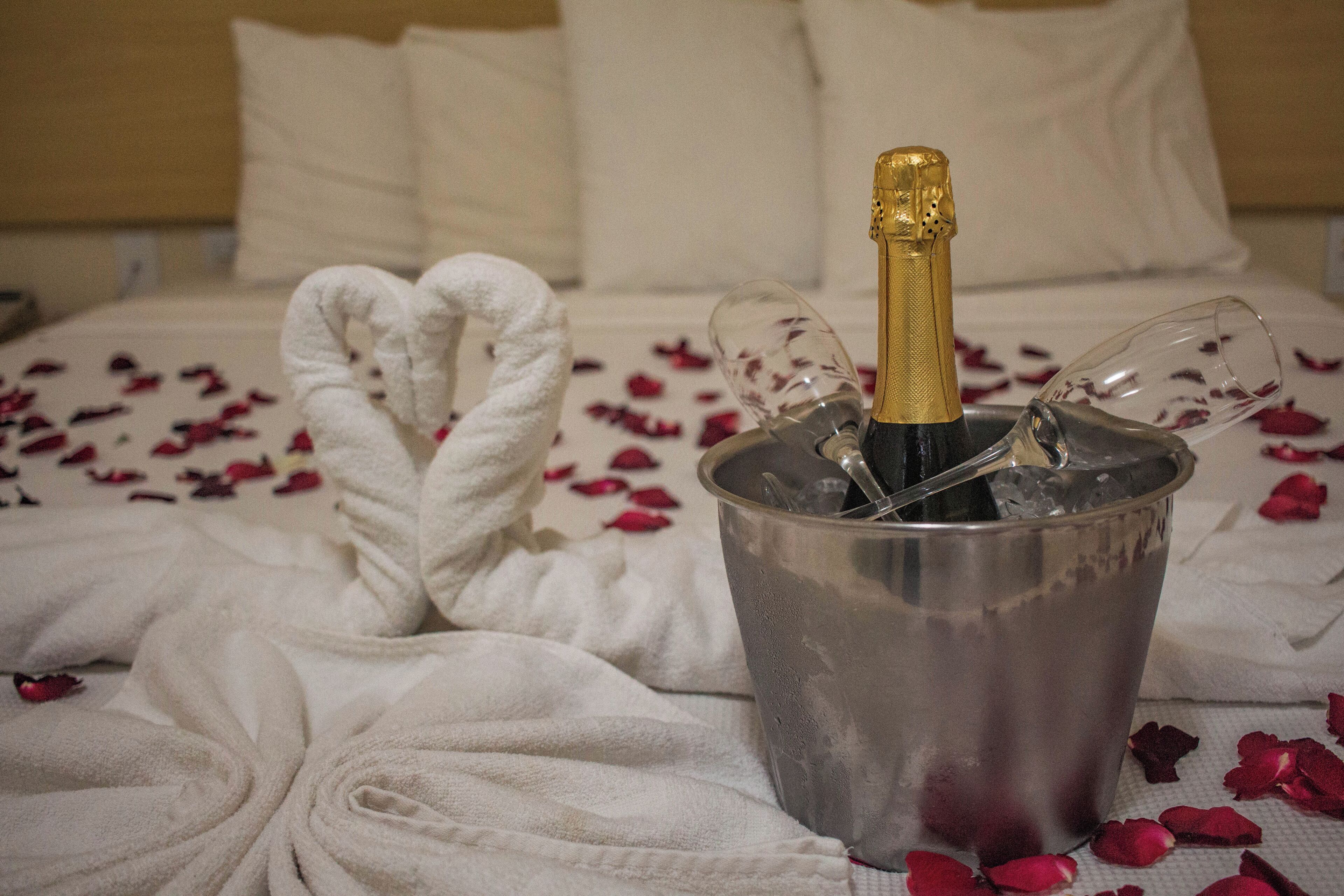 Una cama con una botella de champán en un cubo, rodeada de pétalos de rosa y toallas blancas.