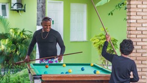 A pool table with balls, a person playing, and a child reaching for a cue stick. - Hotel Parque das Águas (Aracaju)