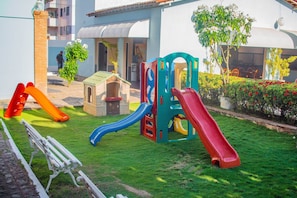 A playground with slides and a small playhouse, surrounded by a well-maintained lawn and a building in the background. - Hotel Parque das Águas (Aracaju)