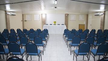 Un salón de conferencias con filas de sillas azules, una mesa blanca con un florero de flores amarillas y paredes de ladrillo.