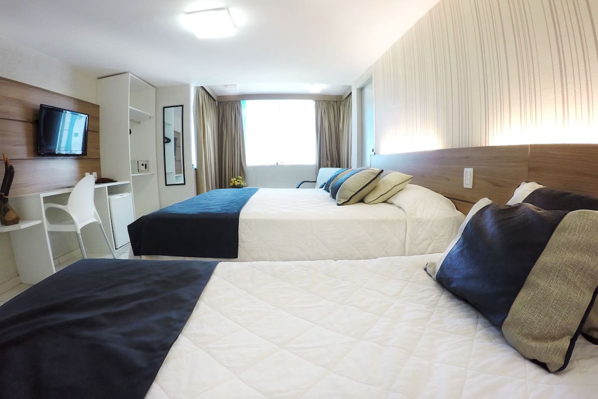 A hotel room with two beds, a desk, a chair, a TV, and a large window with curtains.