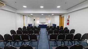 Un salón de conferencias con filas de sillas orientadas hacia una pantalla de presentación, un atril y un extintor en la pared.
