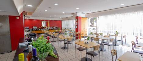 A spacious dining area with multiple tables and chairs, a counter with various items, and large windows providing natural light.