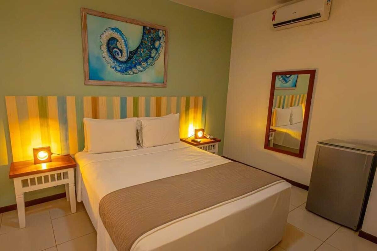 A hotel room with a bed, bedside lamps, a small table, a mirror, and a painting on the wall.