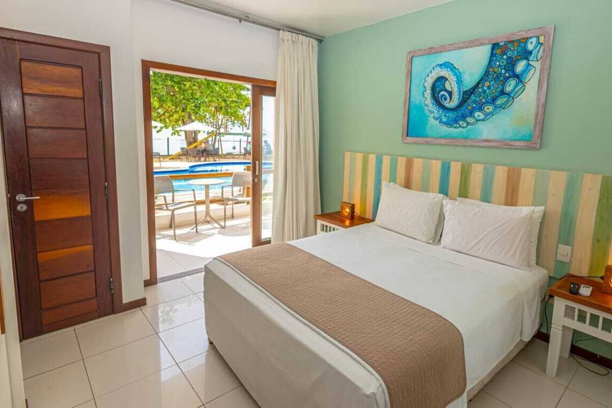 A hotel room with a bed, a painting on the wall, a wooden door, and a view of an outdoor area with tables and chairs.