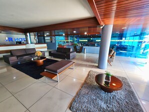 A modern hotel lobby with a reception desk, sofas, a coffee table, and a large pillar. - JR Hotel (João Pessoa)