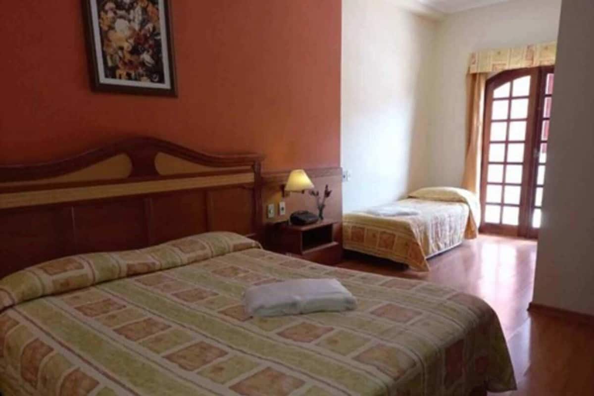 Habitación de hotel con una cama grande, una cama más pequeña, una mesita de noche con lámpara y una ventana con cortinas.