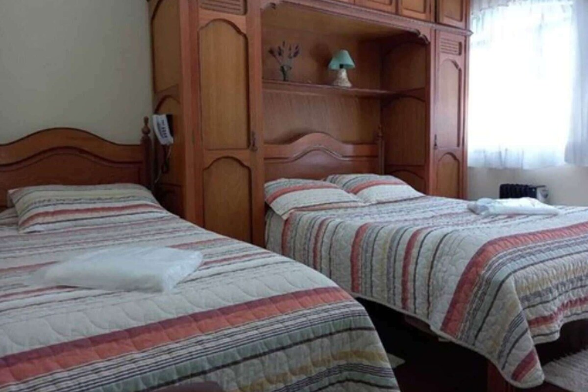 A bedroom with two beds, wooden headboards, a nightstand, a lamp, and a window with curtains.