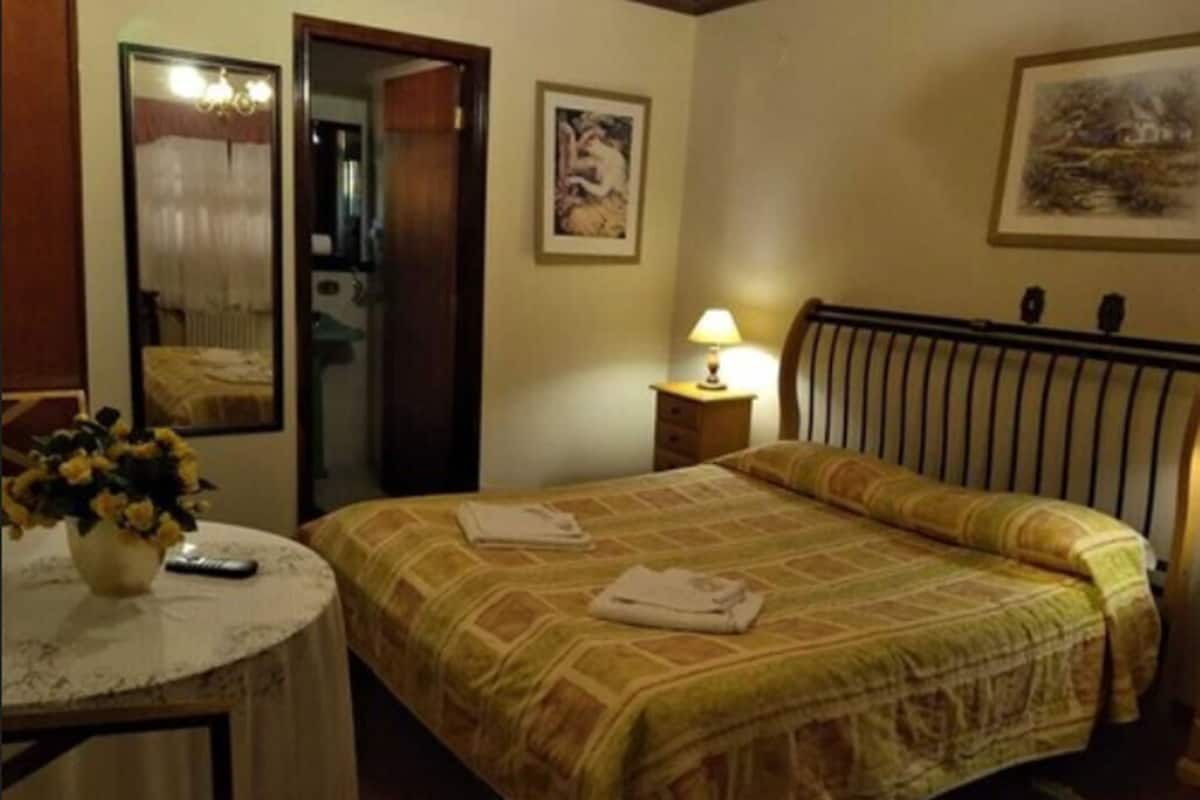 Una habitación de hotel con una cama, una mesita de noche con lámpara, una mesa redonda con un jarrón de flores y un espejo.