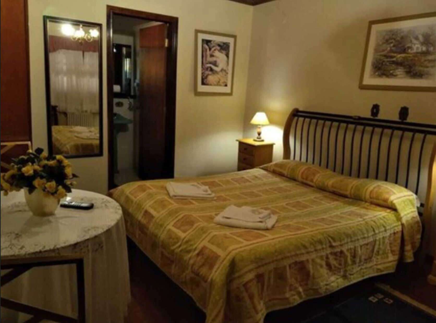 A hotel room with a bed, a bedside table with a lamp, a round table with a vase of flowers, and a mirror.
