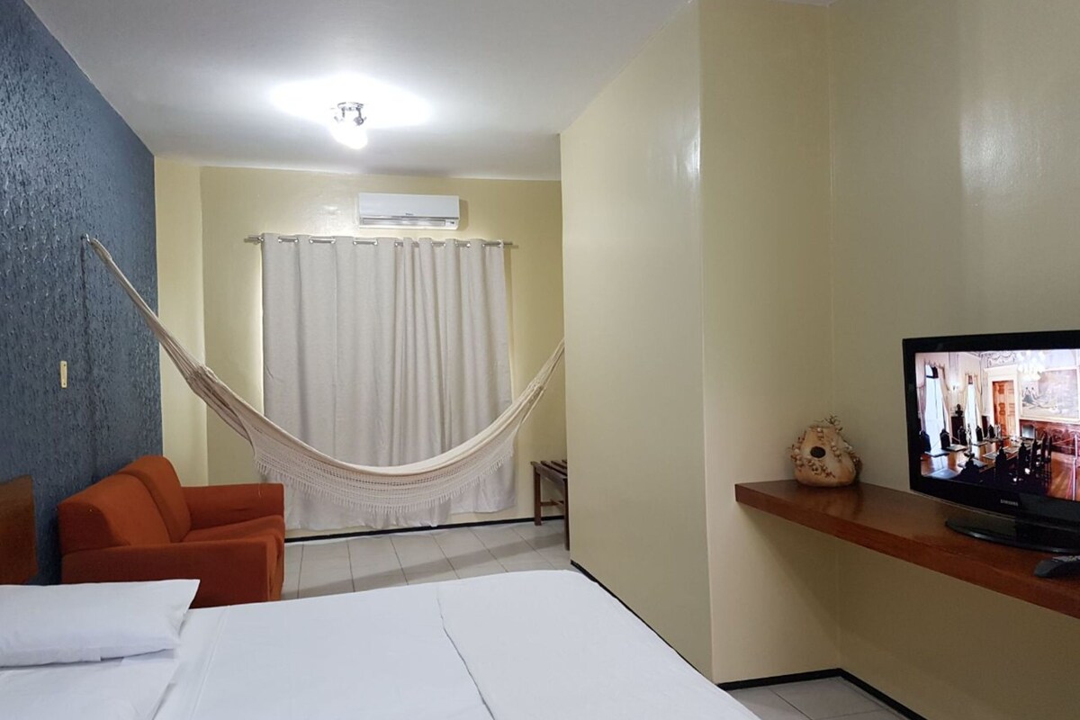 A room with a bed, a hammock, an orange chair, a television, and an air conditioning unit.