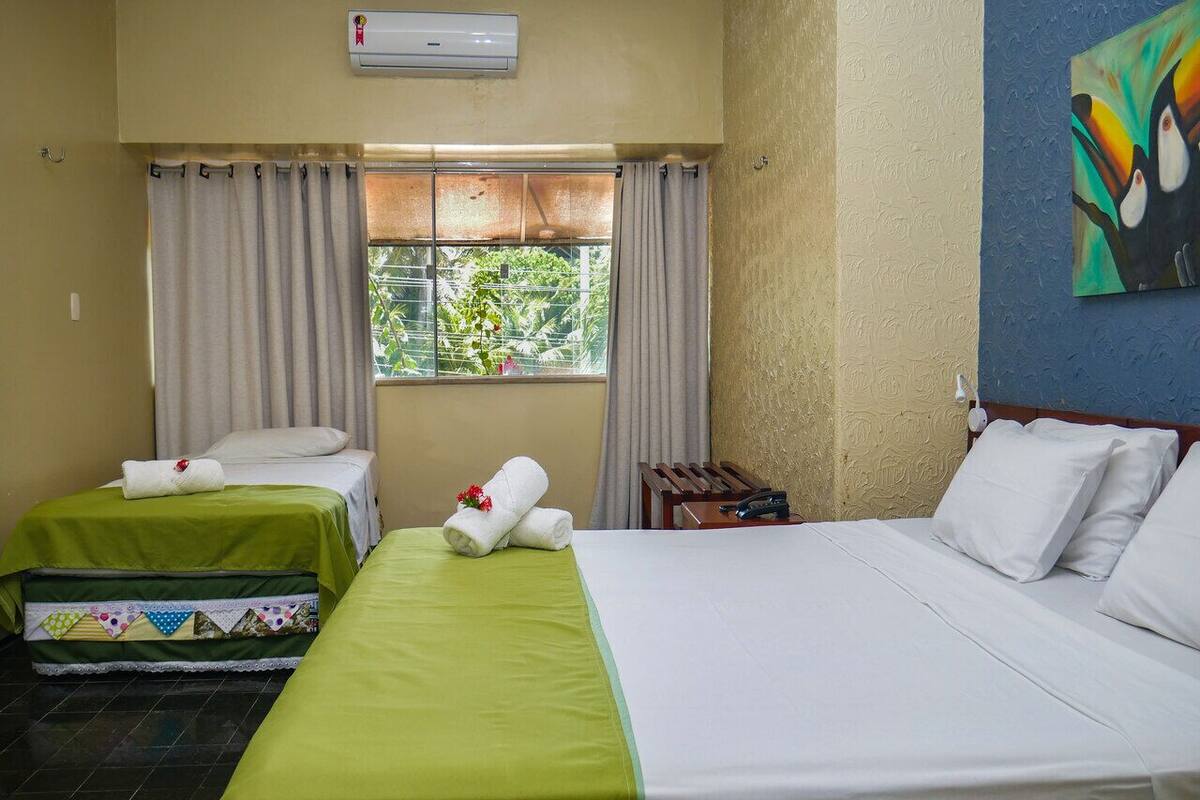 Ein Hotelzimmer mit zwei Einzelbetten, einem großen Bett, einem Fenster mit Vorhängen, einer Klimaanlage und einem Bild an der Wand.
