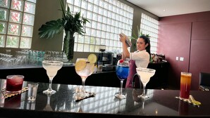 Uma área de bar com um bartender preparando bebidas, uma janela grande com vidro decorativo e diversos copos de coquetel no balcão.