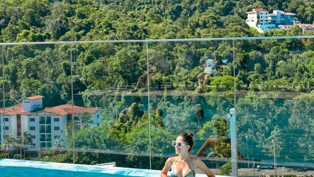 Uma piscina com água azul cristalina, cercada por uma grade, e vista para uma encosta com vegetação densa e edifícios ao fundo.