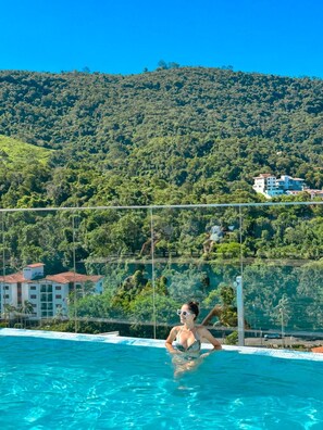 A pool with clear blue water, surrounded by a fence, and a view of a hillside with dense greenery and buildings in the background.
