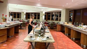 Buffet montado com várias estações de serviço, funcionário atendendo o buffet e área de jantar com mesas e cadeiras.