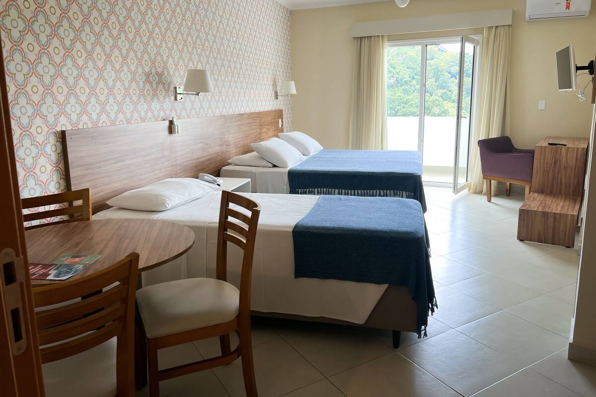 A hotel room with two beds, a desk with a chair, a small table, a ceiling fan, and a window with curtains.