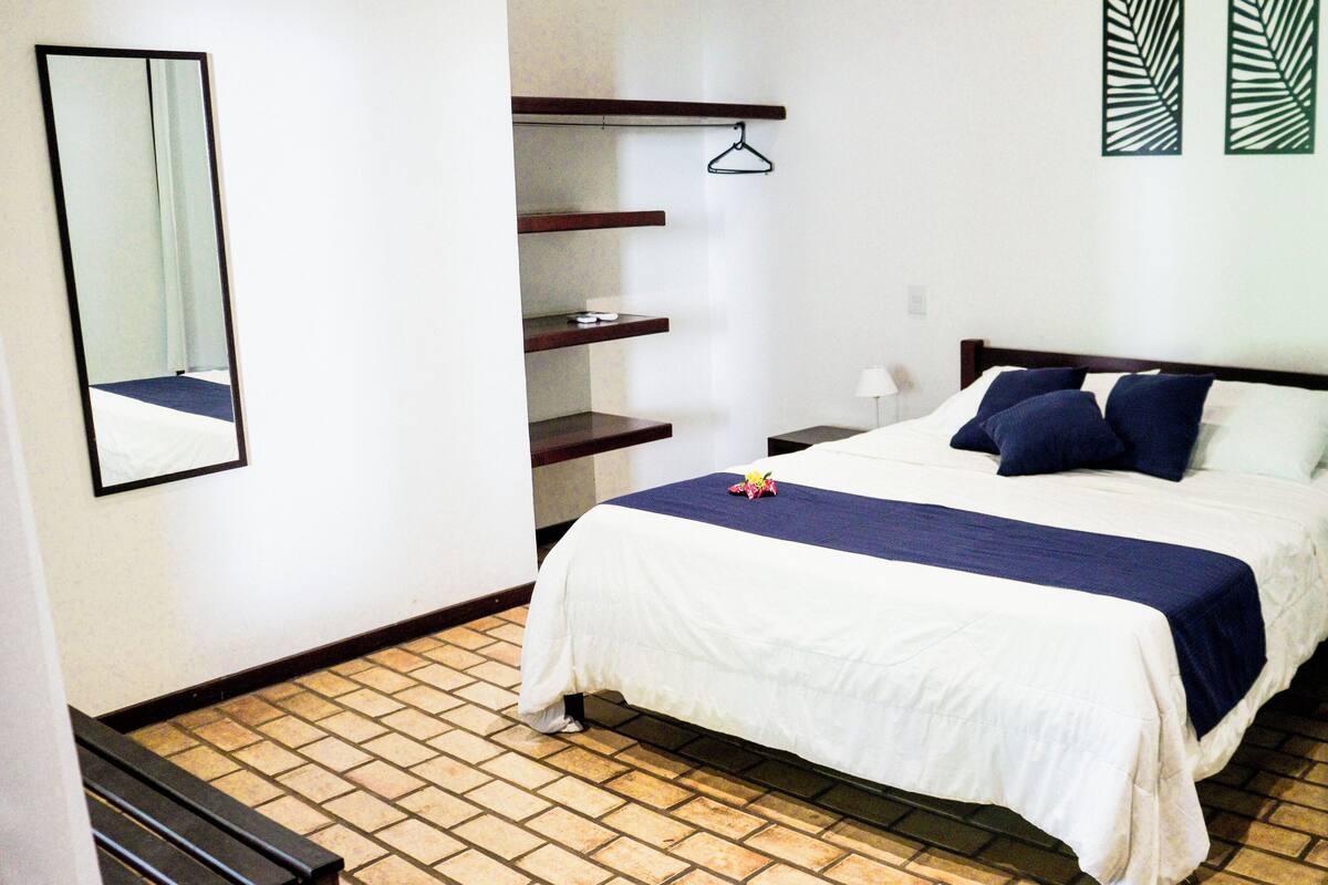 Un dormitorio ordenado con cama, mesitas de noche, una lámpara colgante y un espejo.