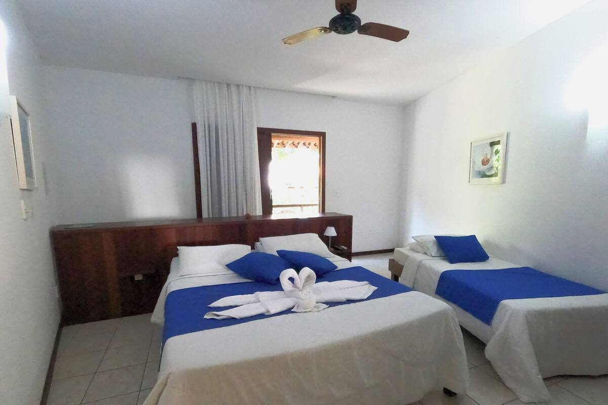 Habitación de hotel con dos camas, cabecera de madera, ventilador de techo, ventana con cortinas y una mesita de noche con lámpara.
