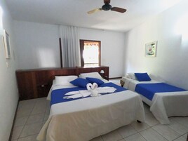 Habitación de hotel con dos camas, cabecera de madera, ventilador de techo, ventana con cortinas y una mesita de noche con lámpara.
