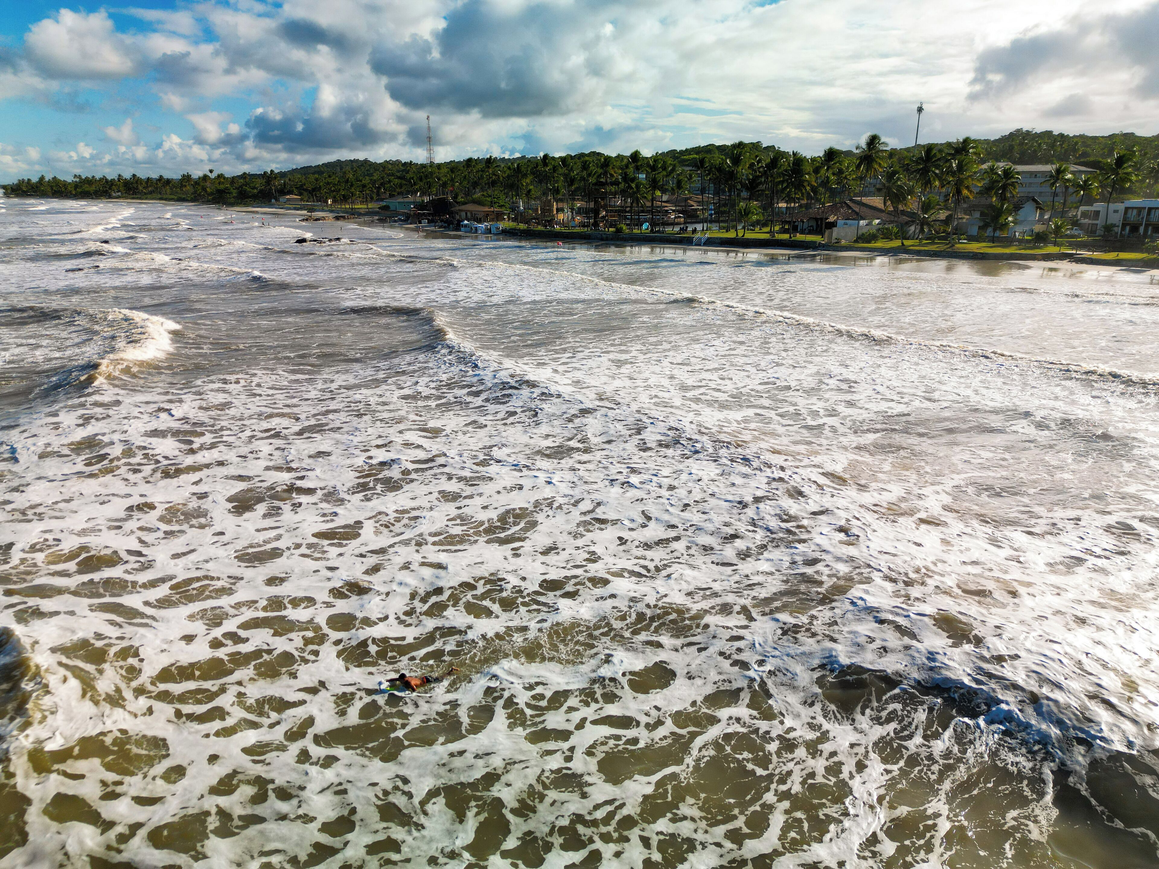 Uma praia com ondas e uma vista distante de uma área residencial com casas e palmeiras.
