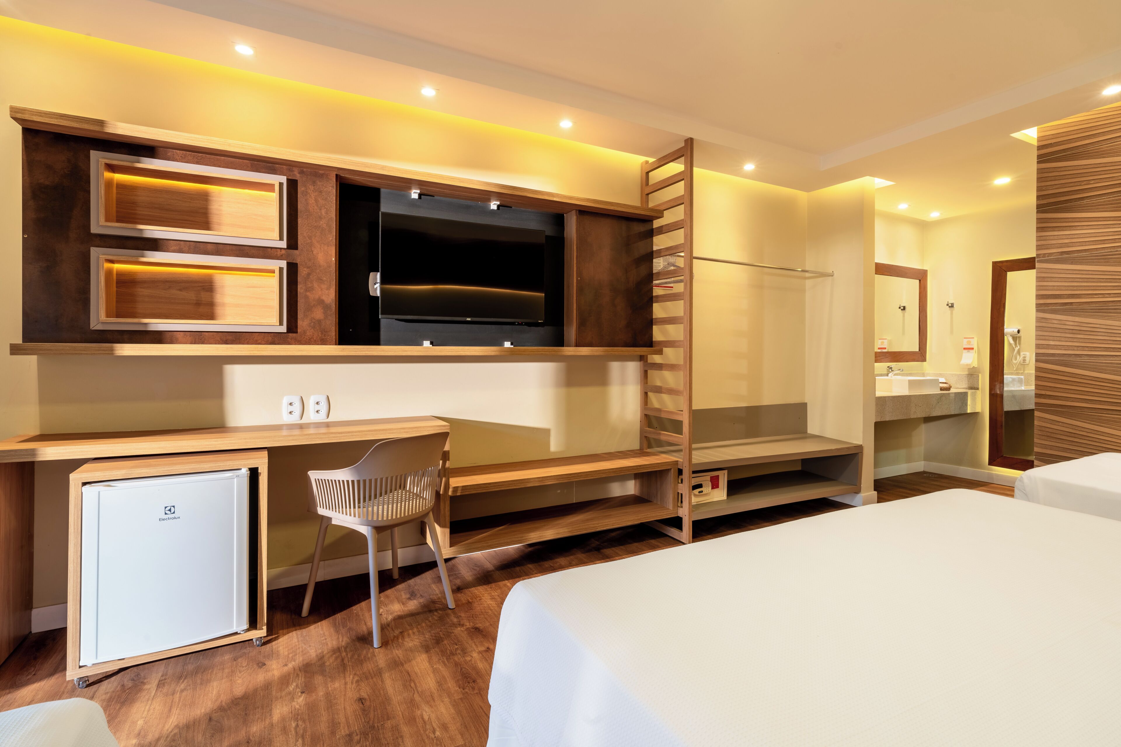 Habitación de hotel con cama, escritorio, silla, televisor, refrigerador pequeño y baño con espejo y lavamanos.