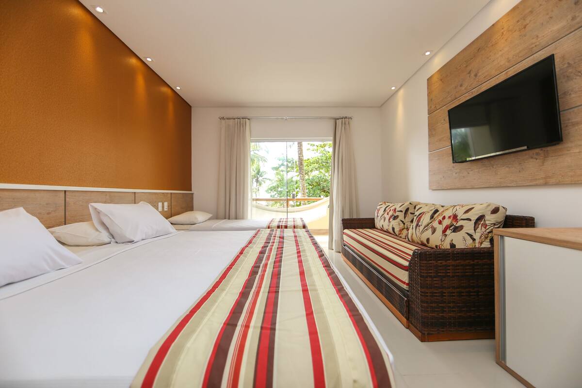 Una habitación de hotel moderna con una cama grande, un sofá, un televisor montado en la pared y un balcón con vistas a zonas verdes.