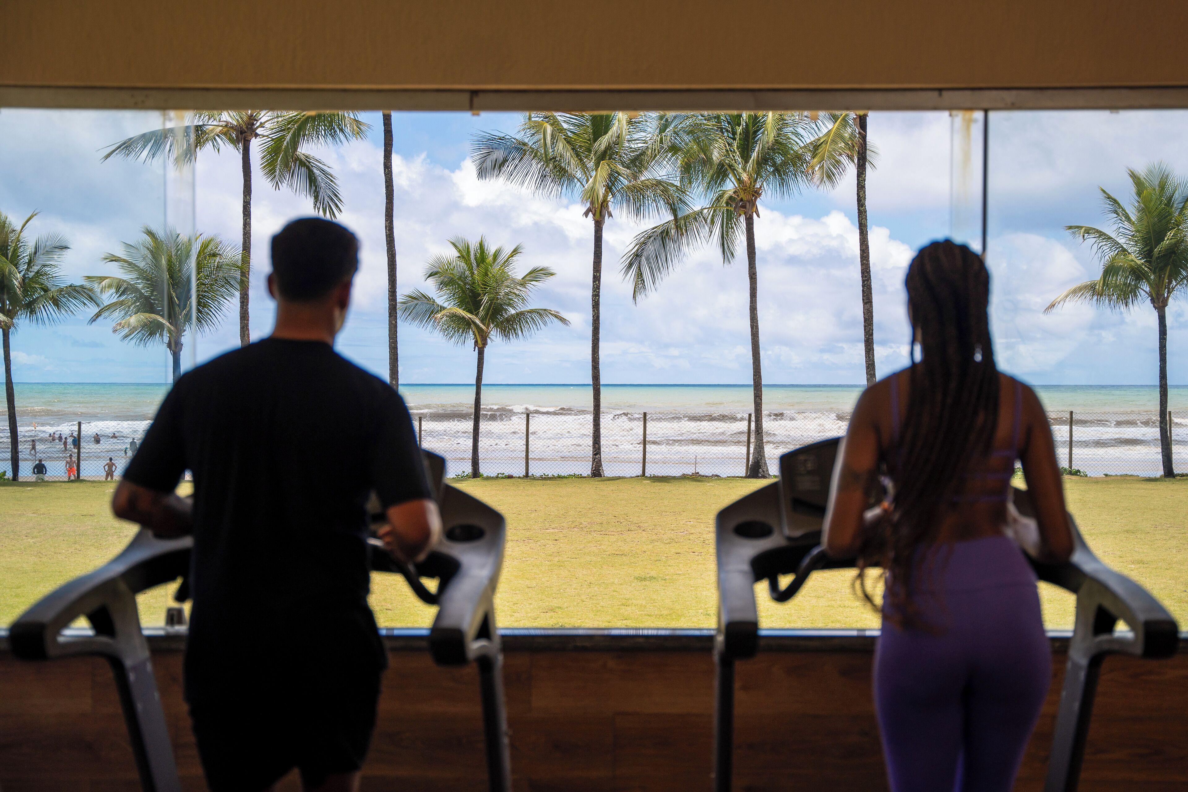 Zwei Personen laufen auf Laufbändern mit Blick auf einen Strand, Palmen und das Meer.