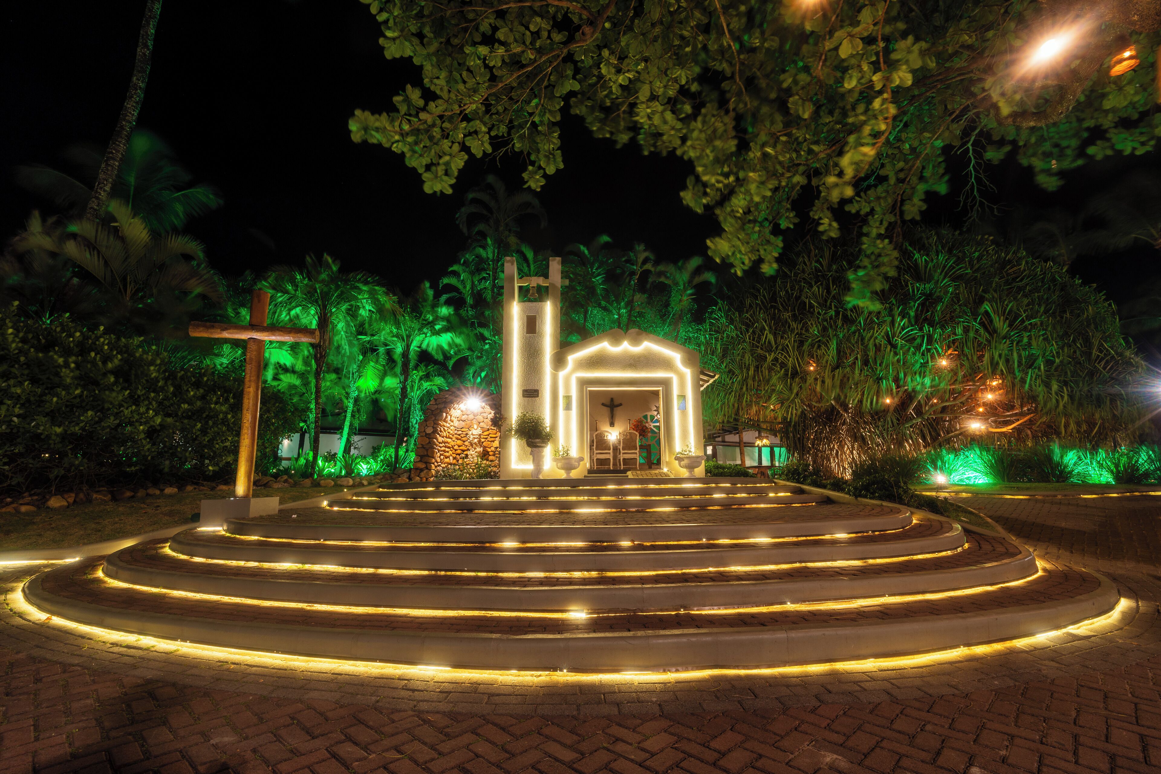 Vista noturna de uma igreja com cruz, escadas que a levam e vegetação ao redor.
