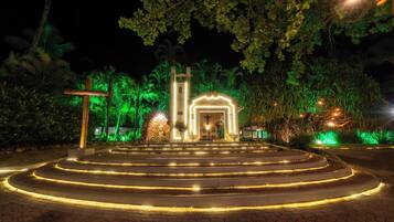 Vista noturna de uma igreja com cruz, escadas que a levam e vegetação ao redor.