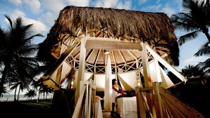 Una estructura de techo de paja con un espacio central abierto, rodeada de palmeras y vista a la playa.