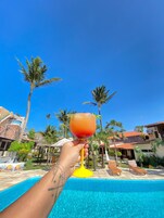 A hand holding a cocktail glass over a swimming pool with palm trees and buildings in the background.