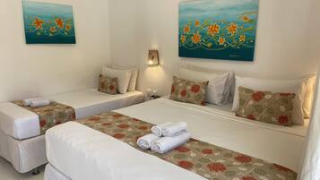 Habitación de hotel con dos camas individuales, una cama grande y una mesita de noche. Se observan cubrecamas y almohadas con estampado floral, además de dos cuadros colgados en la pared.