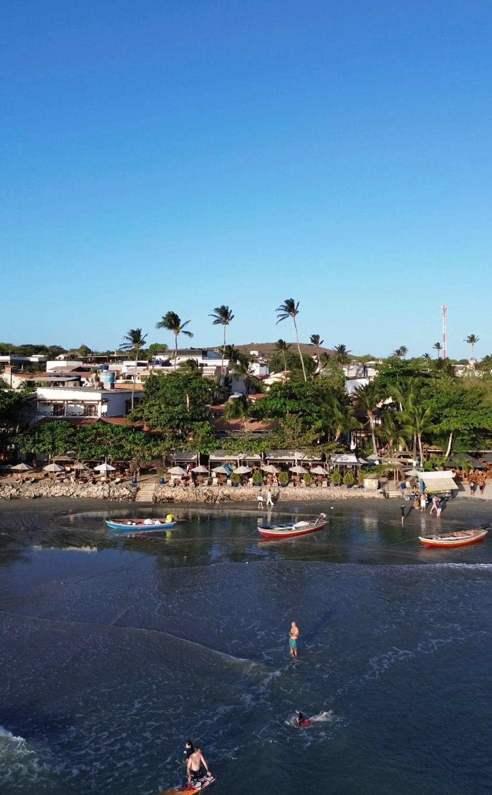 A coastal area with boats on the water, people swimming, and a beach with umbrellas and buildings in the background.