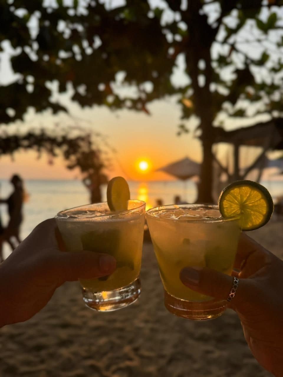 Two glasses of a beverage with a slice of lime, held by hands against a sunset background.