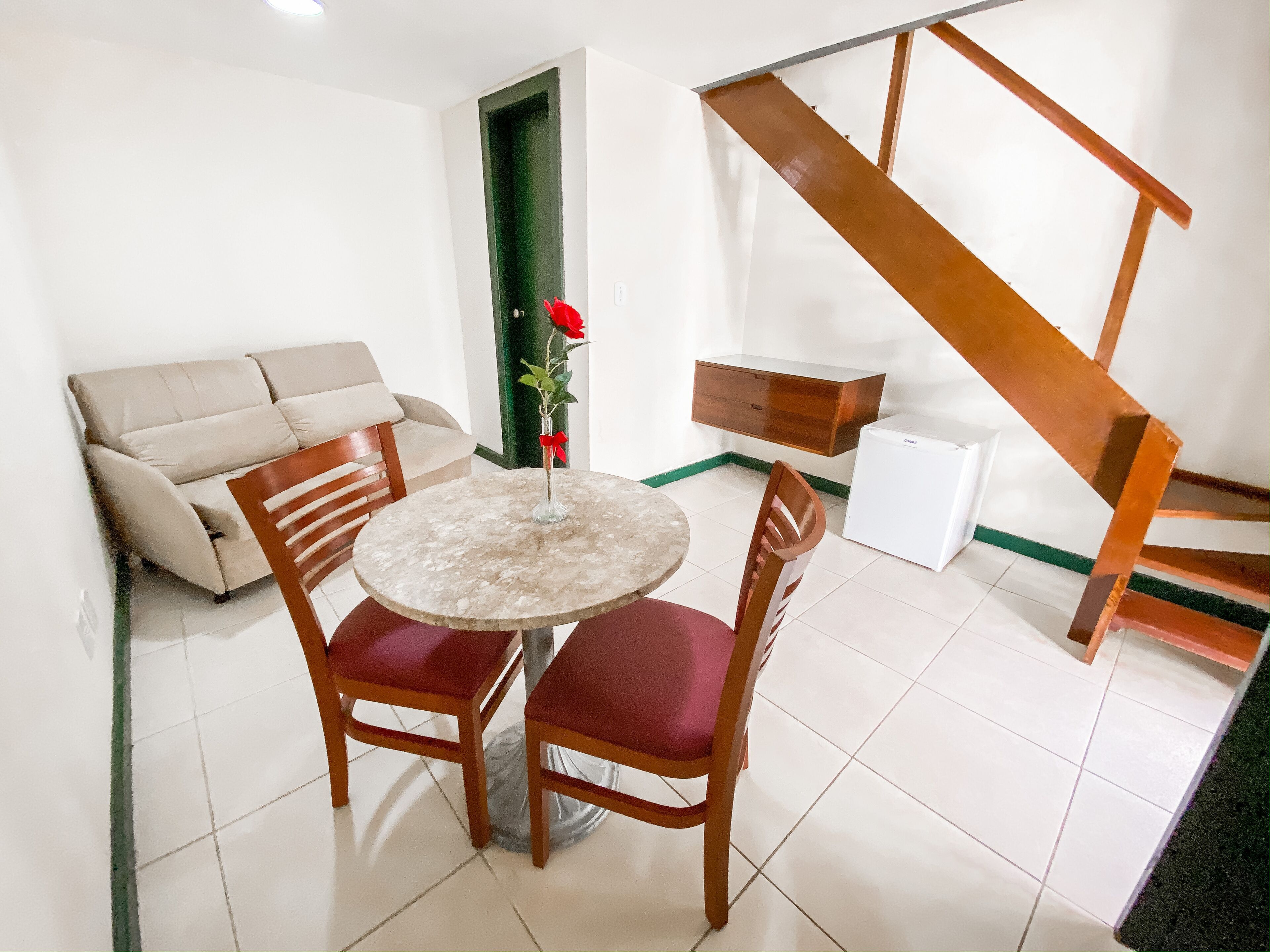 A dining area with a round table, two wooden chairs, a vase with a red flower, a sofa, and a staircase.