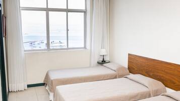 A hotel room with two beds, a window with a view of the sea, and a nightstand with a lamp.