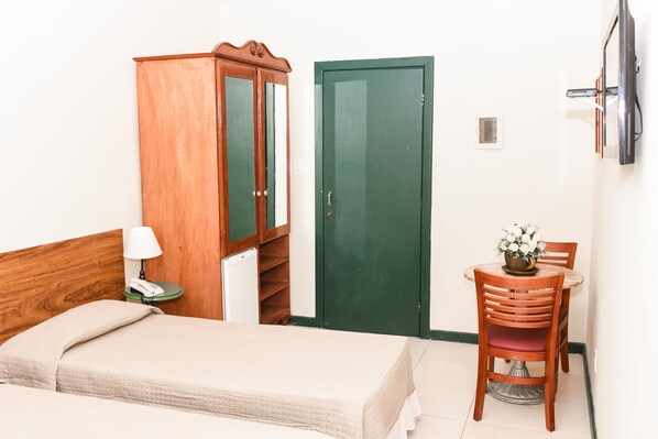 A hotel room with a bed, a wooden wardrobe, a green door, a small table with a chair, and a TV mounted on the wall.