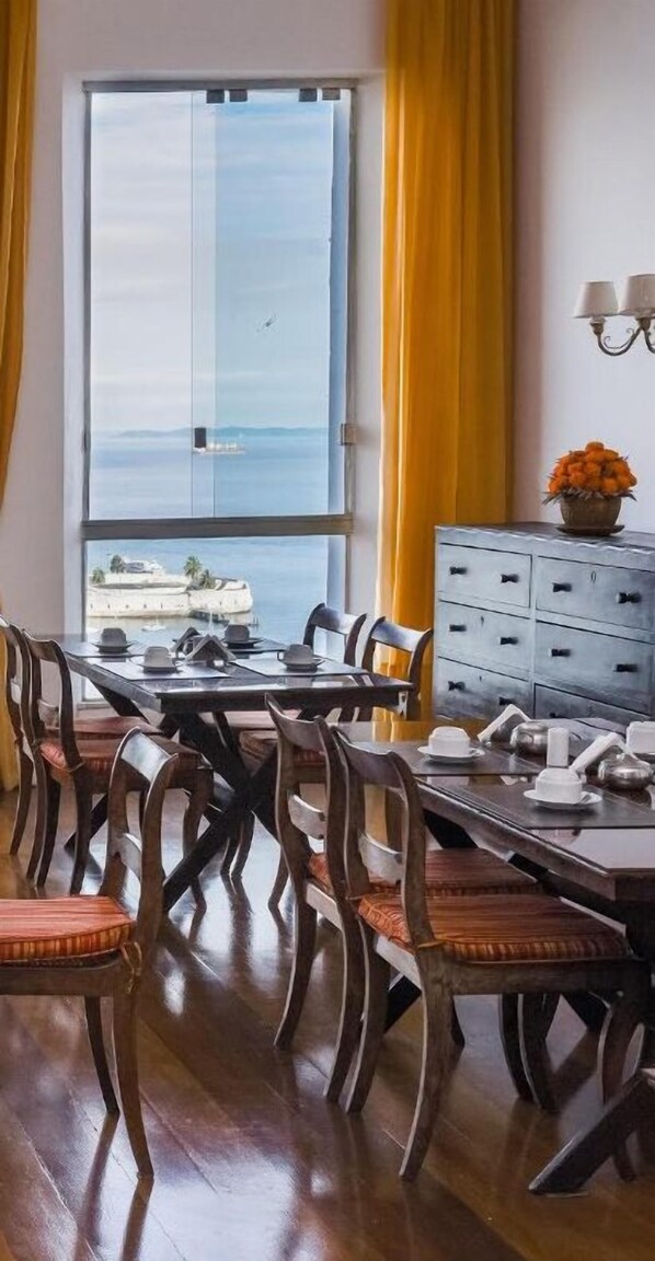 Un comedor con una mesa preparada para cuatro, sillas de madera, un armario con cajones y vistas al mar a través de un ventanal grande.