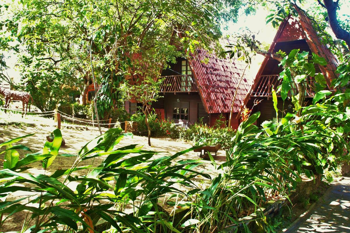 A rustic wooden house with a sloped roof, surrounded by lush greenery and a rope fence.