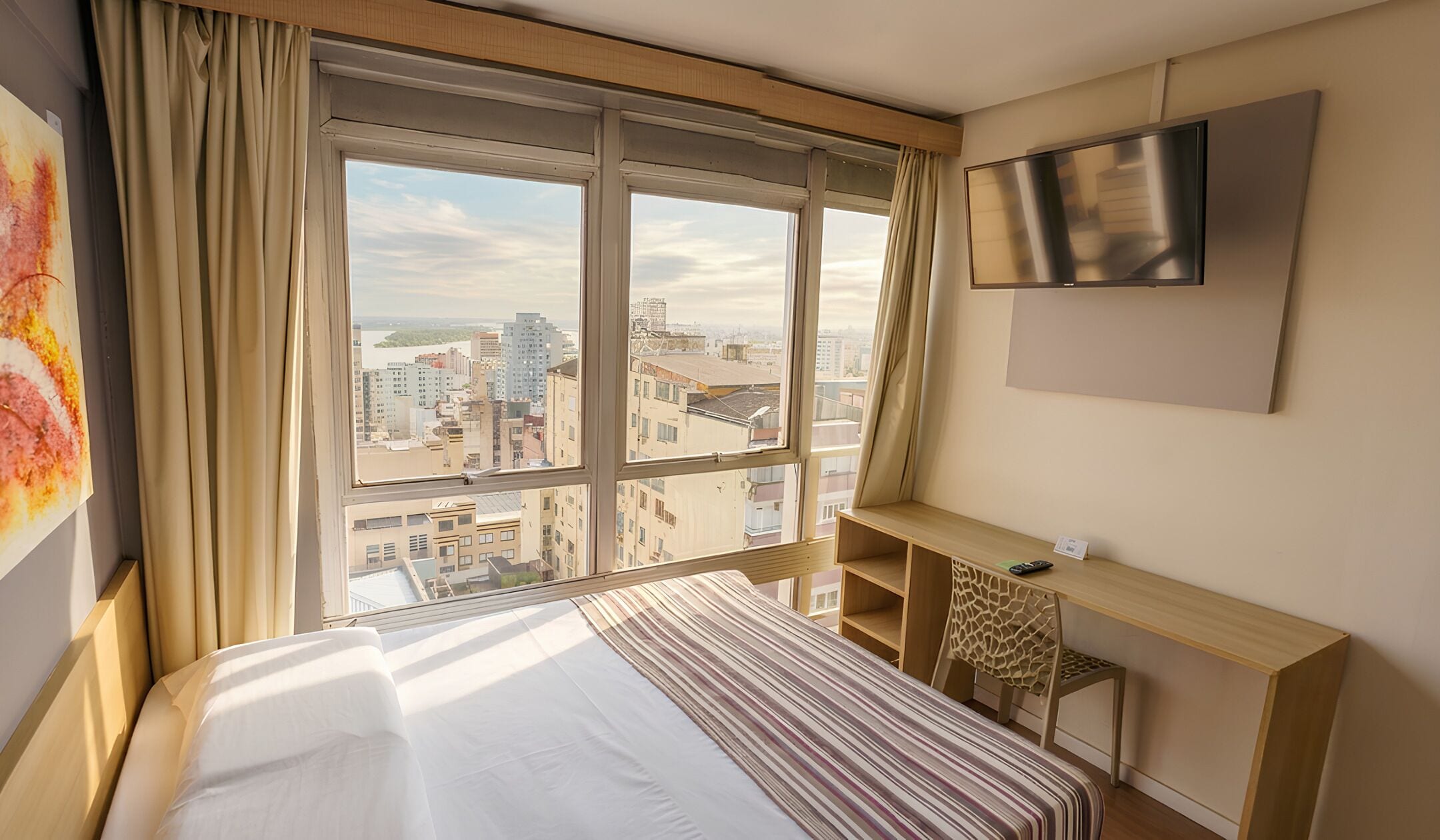 Quarto de hotel com uma janela grande, uma cama, uma escrivaninha e uma televisão.