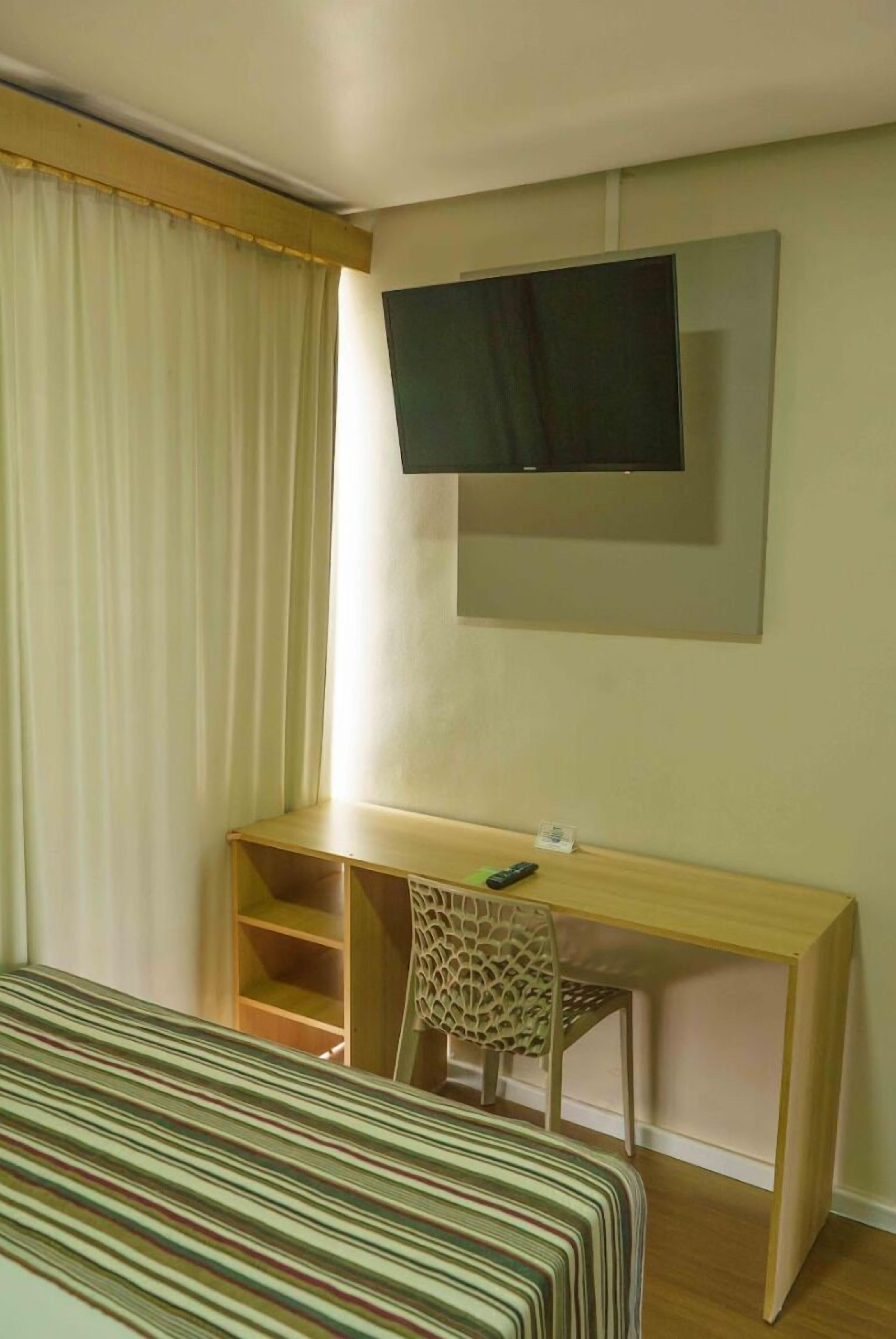A hotel room with a bed, a desk with a chair, a television mounted on the wall, and a window with curtains.