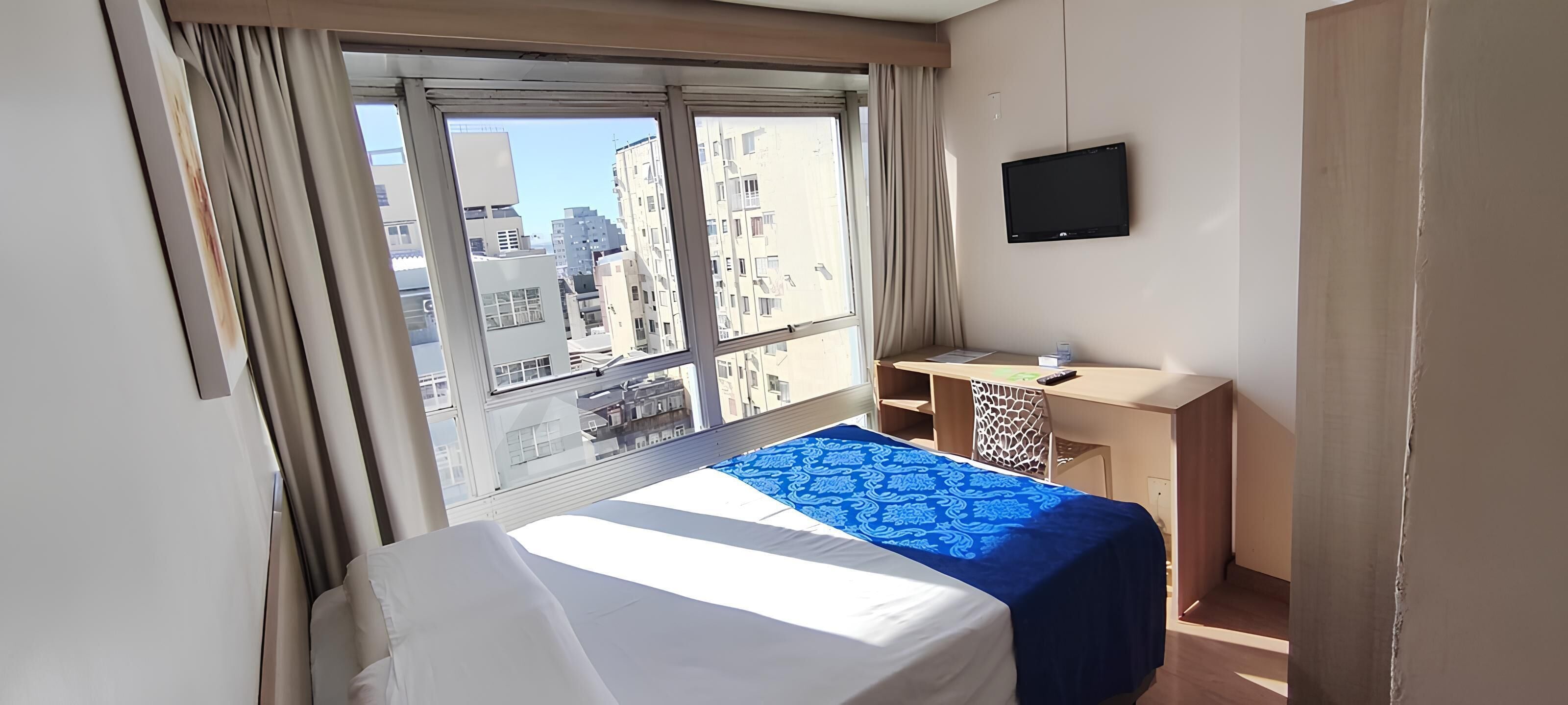 A hotel room with a bed, a desk, a television, and a window with a city view.