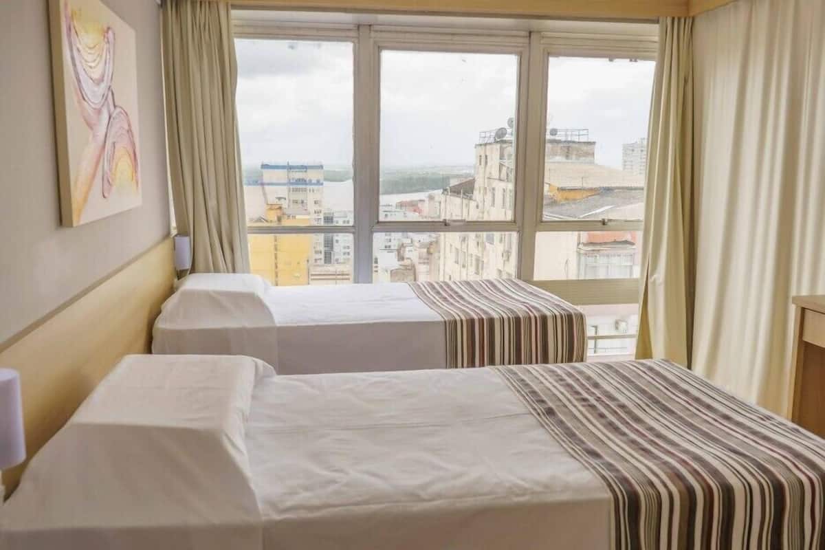 Quarto de hotel com duas camas, janela com vista para a cidade e um quadro na parede.