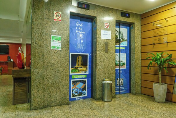 Um elevador com portas azuis, uma lixeira, uma planta em vaso e placas informativas nas paredes.