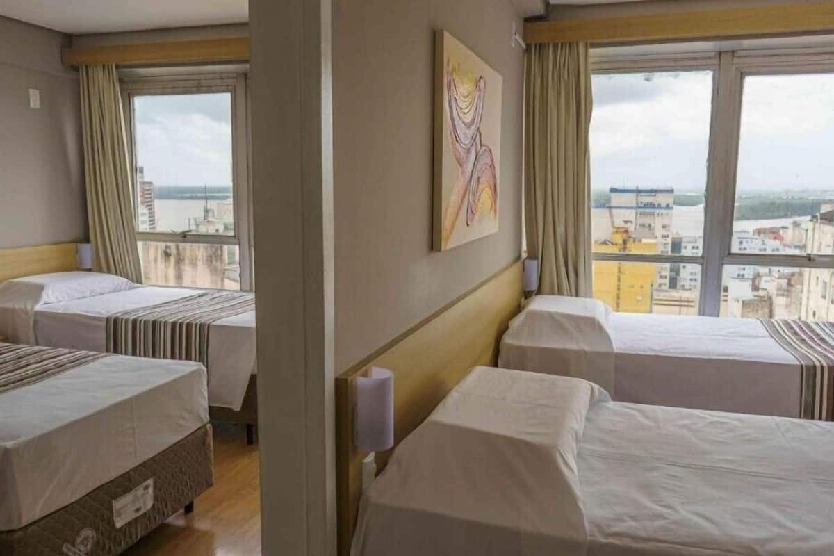 Quarto de hotel com duas camas, um quadro na parede e vista da cidade através de grandes janelas.