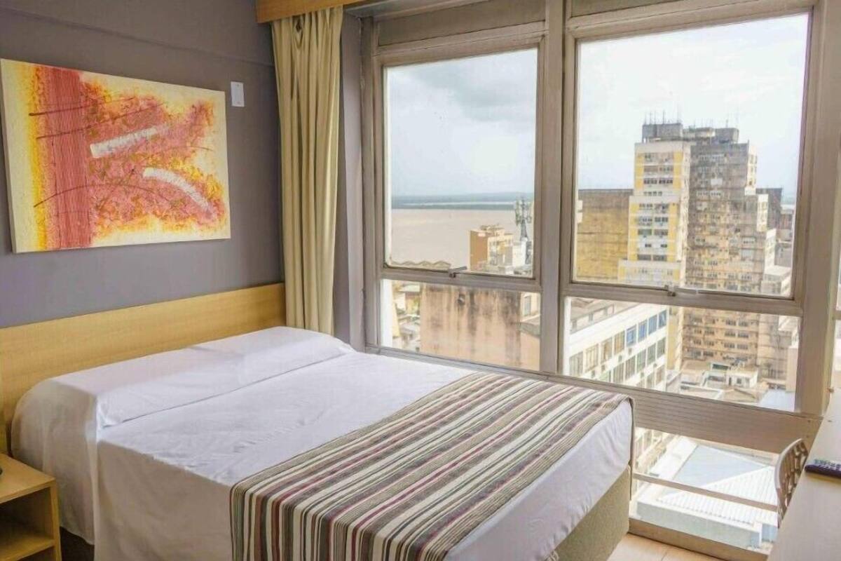 Quarto de hotel com cama, janela com vista para a cidade e uma obra de arte abstrata na parede.
